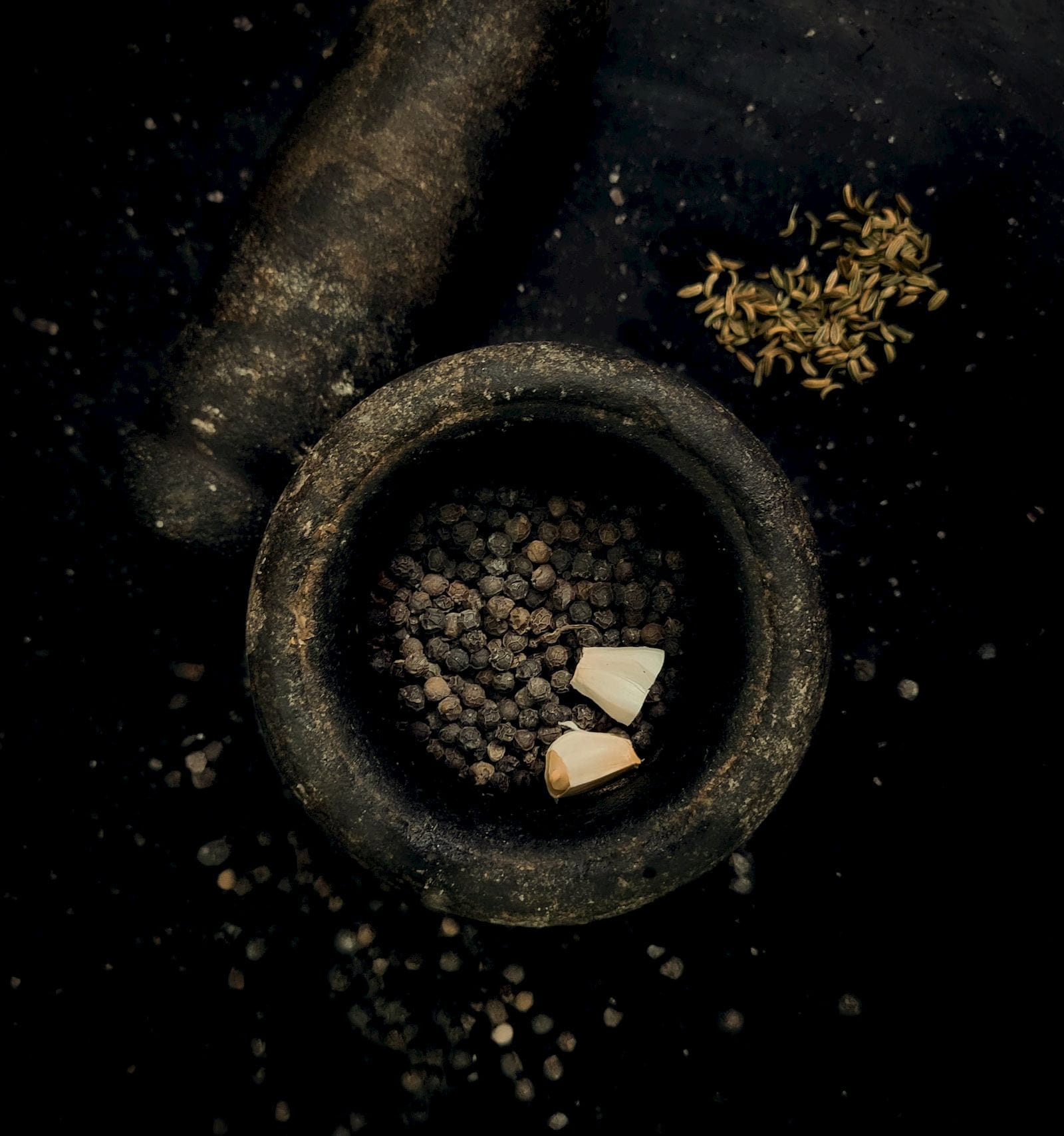 A dark stone mortar holding black peppercorns, with fennel seeds scattered beside it