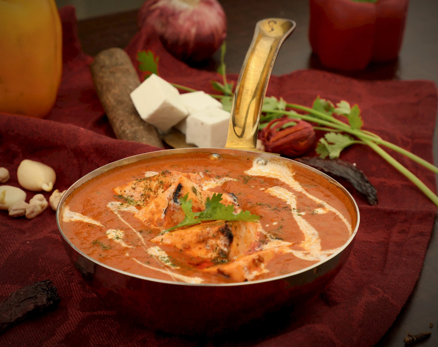 Paneer tikka in a spiced tomato masala in a brass-handled pan, with fresh paneer behind