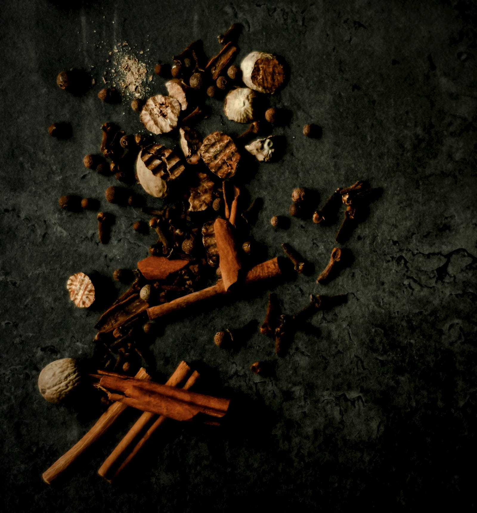 Cinnamon sticks, cloves, nutmeg and mace scattered across dark stone