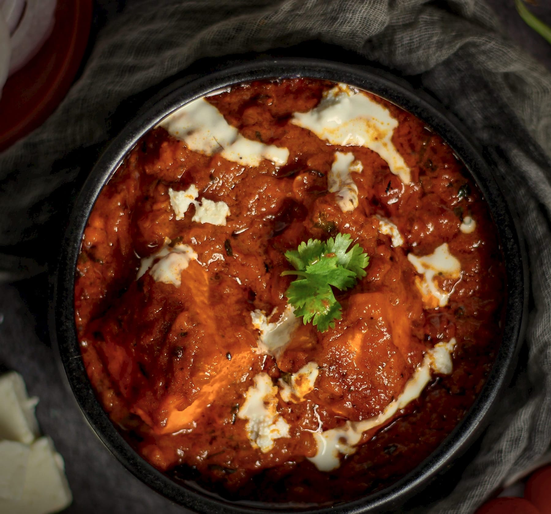 Generous pieces of paneer in a deep, coarse masala in a black ceramic bowl, with fresh paneer beside it